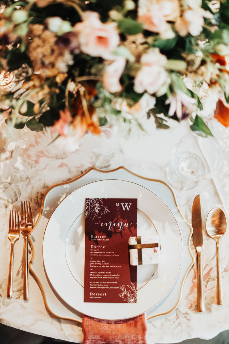 Wedding reception table with a plate, charger, gold flatware and burgundy wedding menu and macaron box