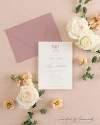 The Ribbon Bow dusty rose shower invitation with a hand-illustrated bow, beside its mauve envelope, styled with white and peach roses