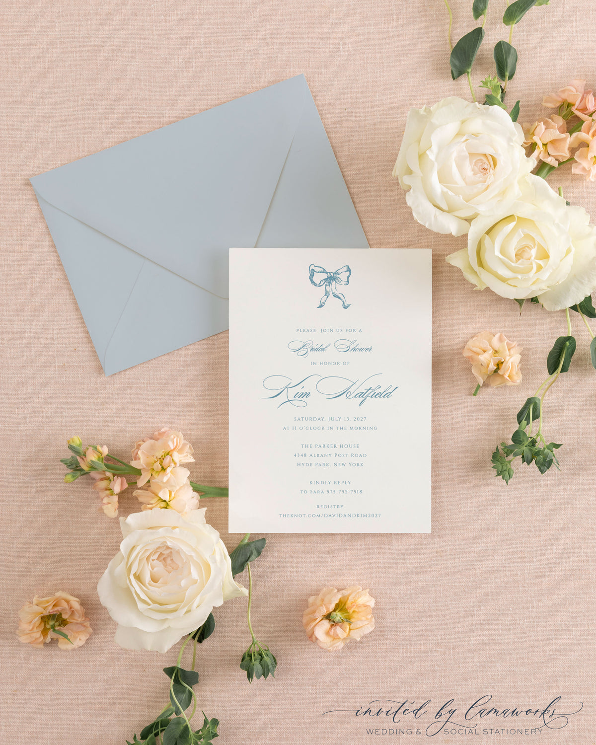 The Ribbon Bow dusty blue shower invitation with a hand-illustrated bow, beside its dusty blue envelope, styled with white and peach roses