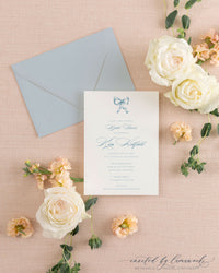 The Ribbon Bow dusty blue shower invitation with a hand-illustrated bow, beside its dusty blue envelope, styled with white and peach roses