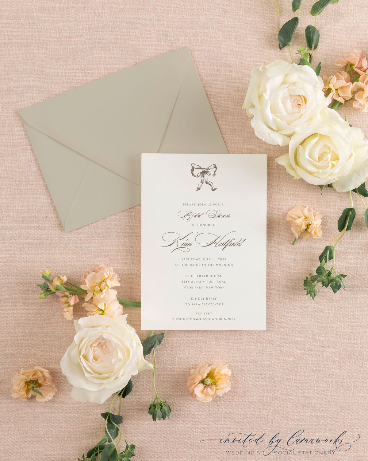 The Ribbon Bow taupe shower invitation with a hand-illustrated bow, beside its taupe envelope, styled with white and peach roses
