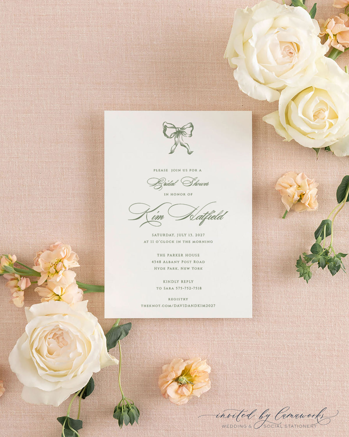 The Ribbon Bow sage green shower invitation with a hand-illustrated bow, beside its cream envelope, styled with white and peach roses
