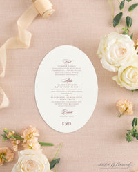 The Kim collection oval die cut wedding menu card in burgundy ink with calligraphy course headers and a K and D monogram, styled with peach ribbon and roses