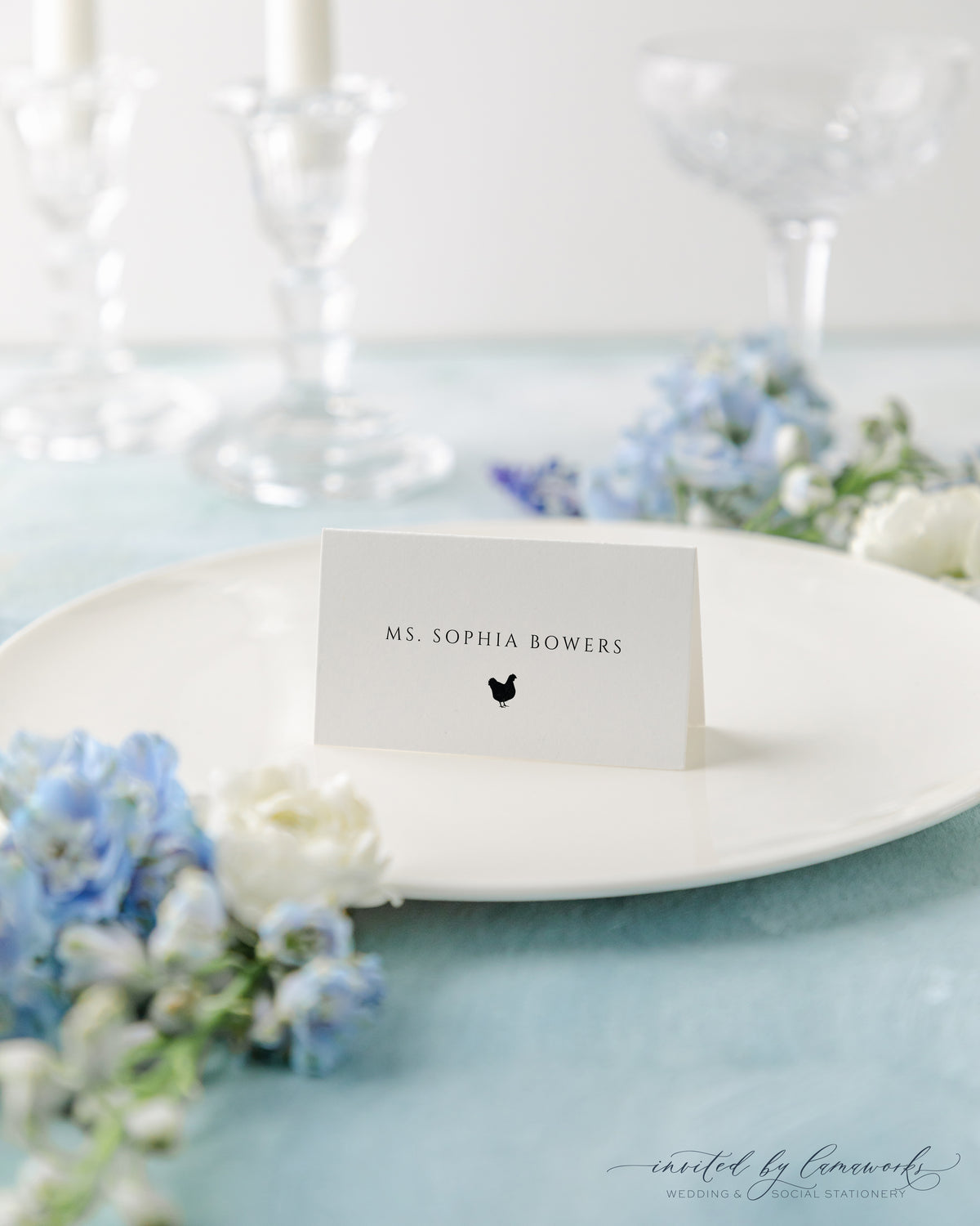 The Kim collection wedding place card with a guest's name and a small meal choice icon, set on a white plate with crystal glassware and florals