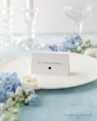 The Kim collection wedding place card with a guest's name and a small meal choice icon, set on a white plate with crystal glassware and florals