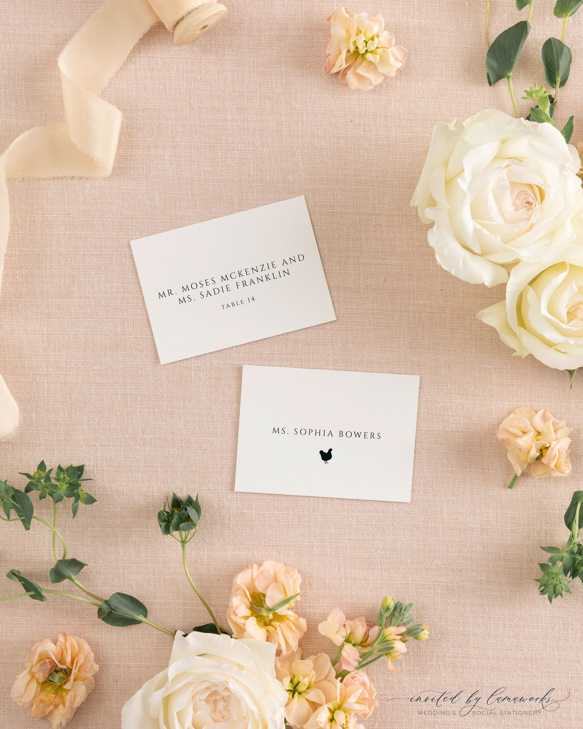 The Kim collection place and escort cards on pink linen with peach ribbon and roses, showing a guest name with meal icon and a couple's names with table number