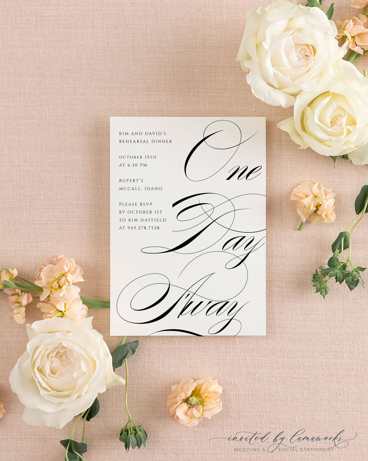 The Kim collection rehearsal dinner invitation with bold black calligraphy reading One Day Away, beside its cream envelope, styled with white and peach roses